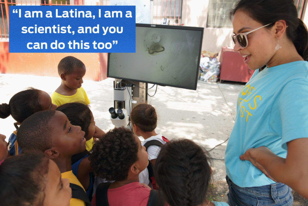 A young Latina woman stands with her hands on her hips smiling at an attentive group of mostly Black children. They are all using a microscope together outside.